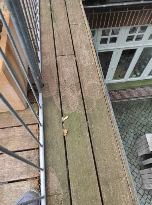 Blick entlang eines Balkonbodens aus Holz mit sichtbaren Verwitterungsspuren, grünlichem Algenbewuchs und Verschmutzungen. Vor der Anwendung des Anti-Belag-Reinigers.
