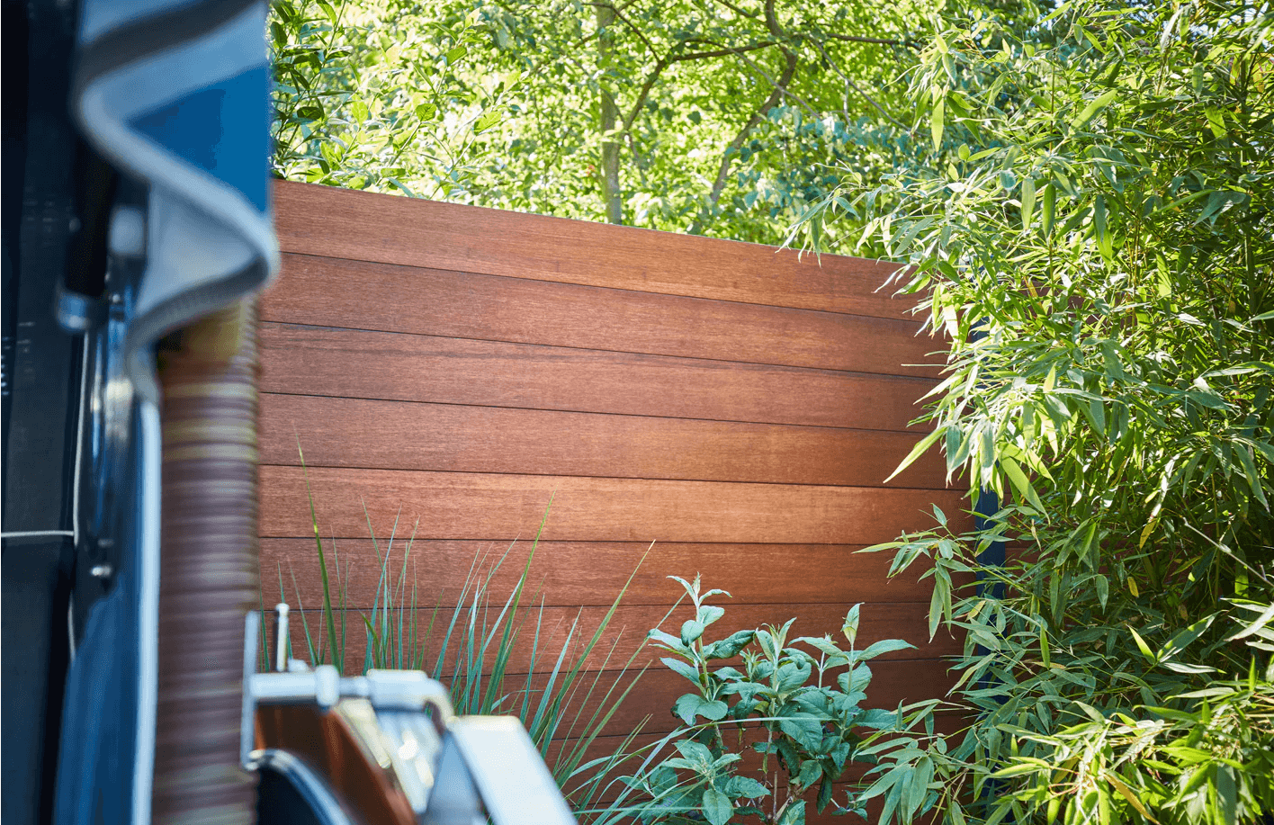 aMbooo Bambus-Sichtschutz Delxue im Garten, umgeben von grünem Bambus und Bäumen, mit einem angeschnittenen Strandkorb im Vordergrund.