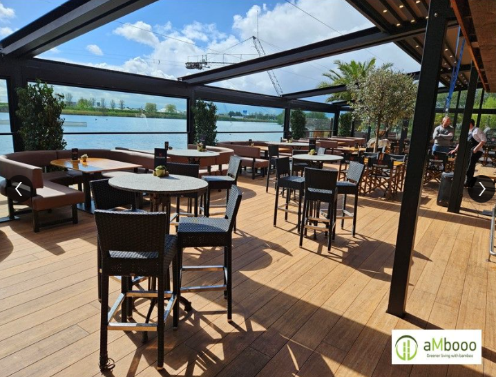 Überdachte Restaurantterrasse mit holzoptischem Terrassenbelag aus Closed Decking Dielen in warmen Sandtönen. Auf der Terrasse stehen zahlreiche Tische und schwarze Flechtstühle. Große Glasfronten und offene Seiten bieten einen weiten Blick auf die Umgebung. Das Sonnenlicht betont die natürliche Holzstruktur des Bodenbelags und schafft eine einladende Atmosphäre.