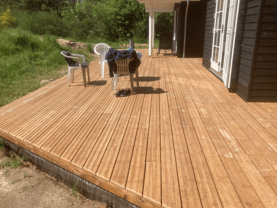 Eine frisch verlegt ambooo Bambus Terrasse mit der Diele Campoverde vor einen Haus mit dunkler Holzfassade, möbliert mit Gartetisch und Stühlen in der Farbe weiß umgeben von grün.