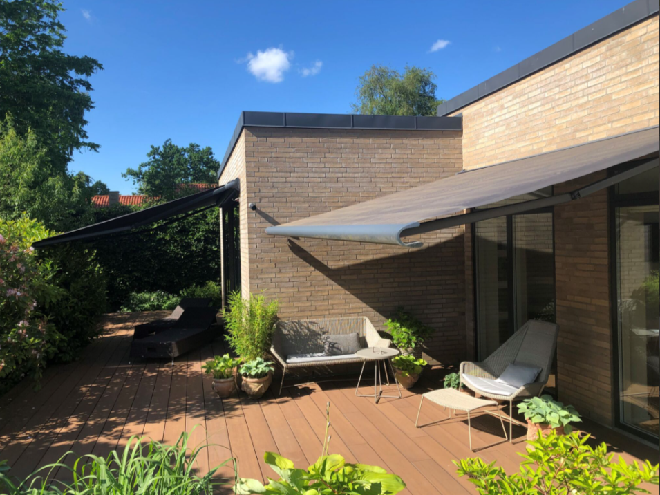 Moderne Terrasse mit Bambus-Terrassendielen Primaverde in warmem Braunton, ausgestattet mit Markisen, Loungemöbeln und Topfpflanzen vor einem Flachdachhaus aus Klinker bei Sonnenschein.