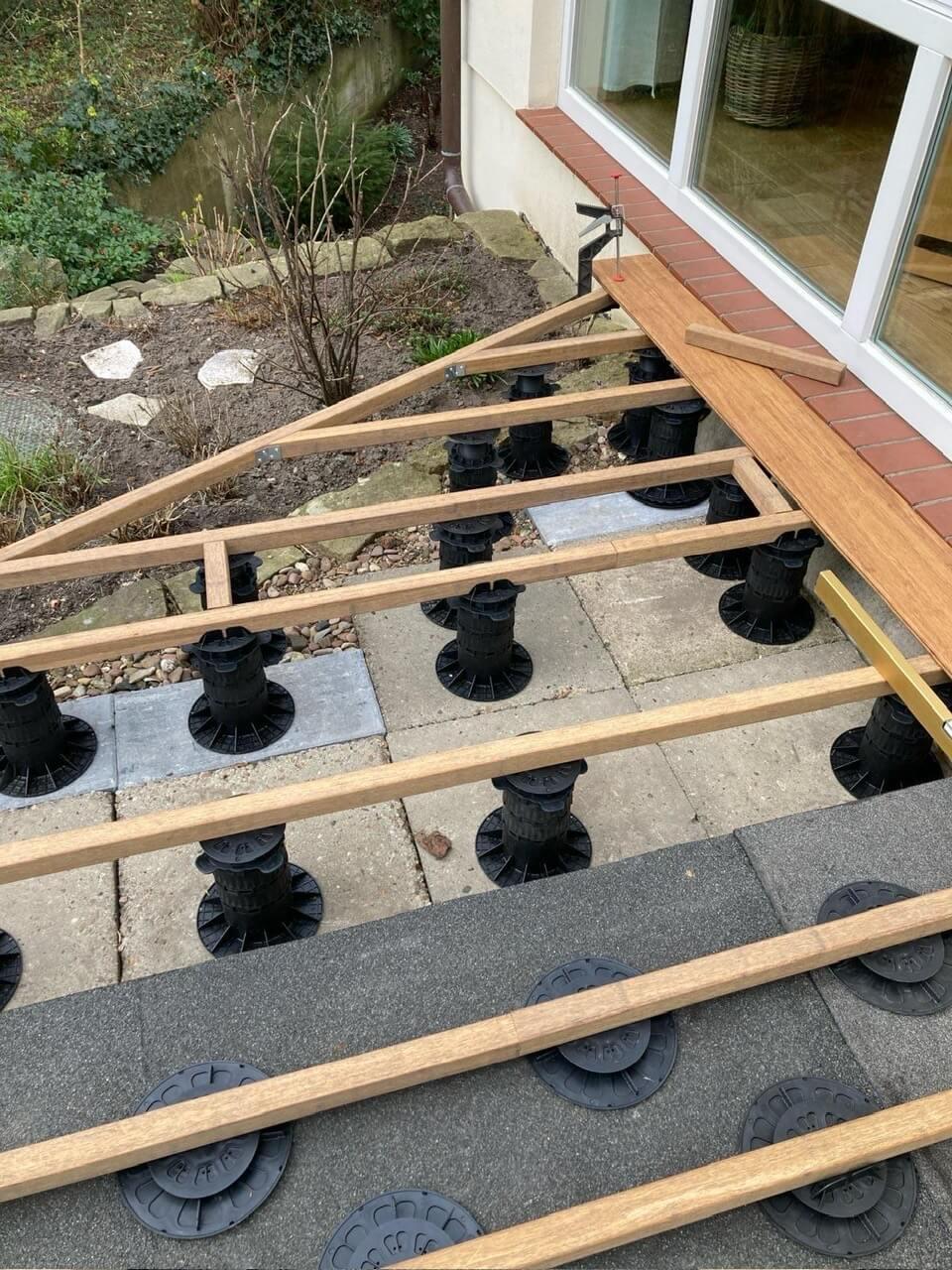 Terrassenunterkonstruktion aus Bambusbalken auf höhenverstellbaren Stelzlagern vor einem Wohnhaus während der Montage.