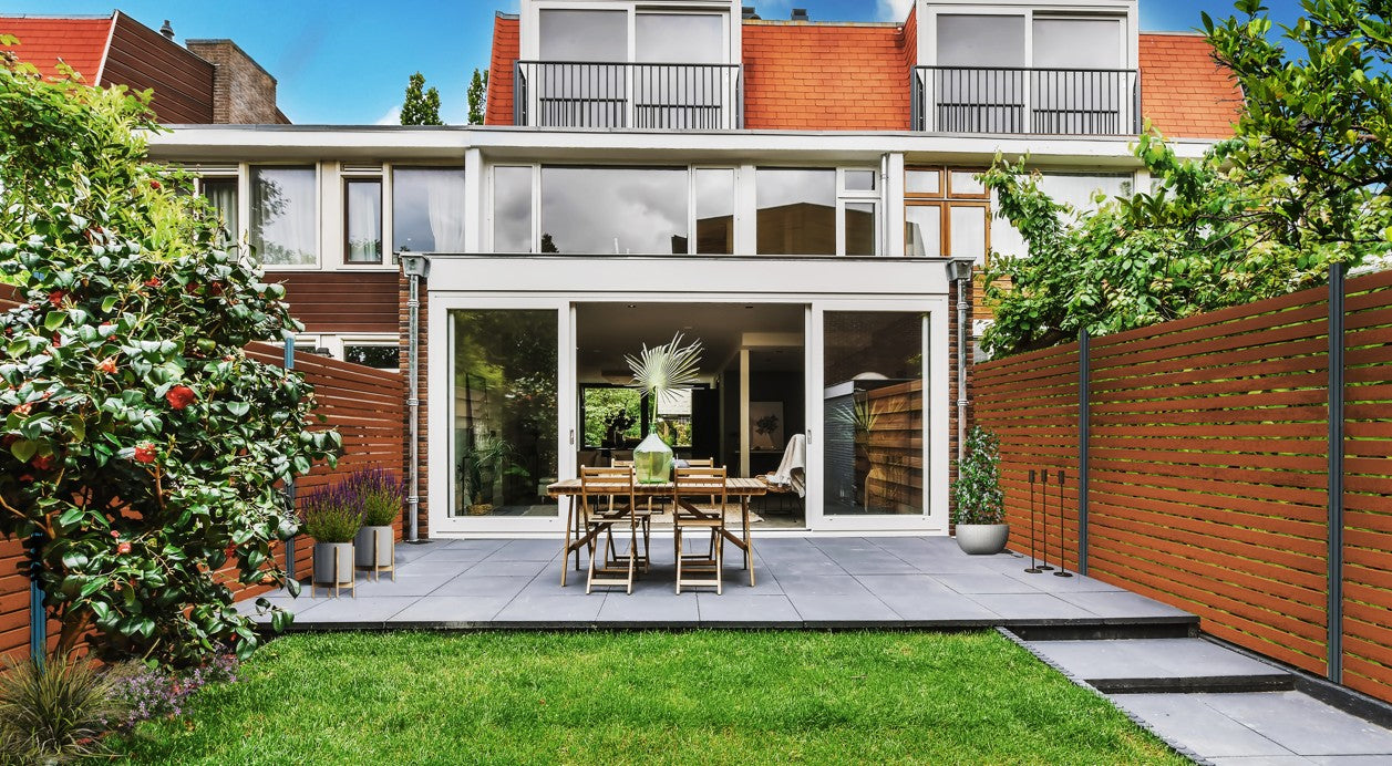 Blick in einen modernen Hinterhof: Eine geflieste Terrasse mit Holztisch und -stühlen vor einer großen Glasfront. Rechts und im Hintergrund begrenzt ein hoher Bambus Sichtschutzzaun mit horizontalen, braunen Latten den Garten. Davor liegt eine gepflegte Rasenfläche, umgeben von grünen Büschen und Pflanzen.