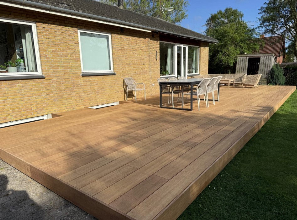 großzügige Terrasse erstellt mit ambooo closed decking Terrassendielen in natürlicher Holzoptik, aussgestattet mit moderenen Essbereich und Loungemöbeln, vor einem Einfamilienhaus mit gelber Klinkerfassade.