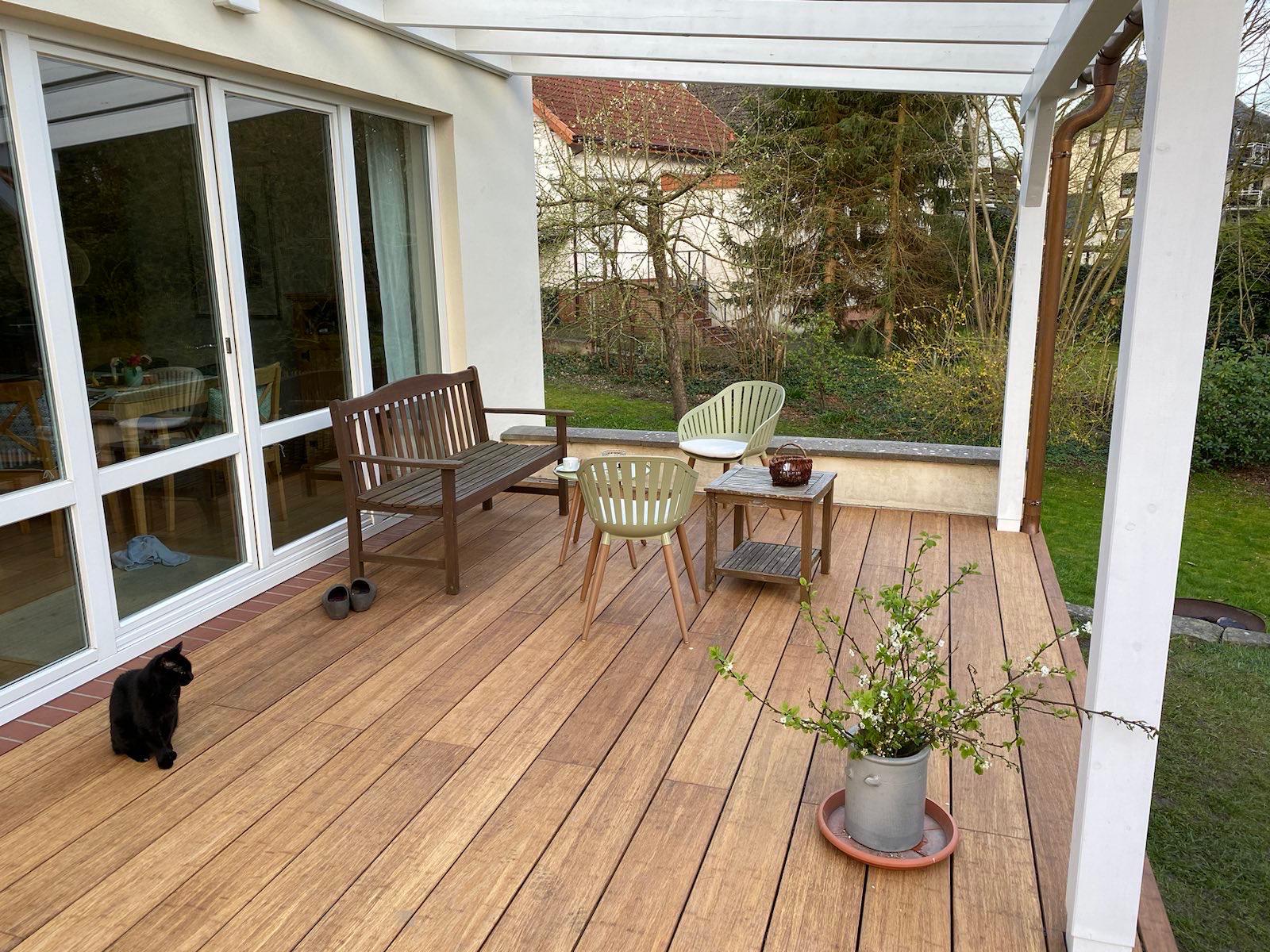 Moderne Terrasse mit hochwertigen glatten XL Bambus Terrassendielen, in der Farbe Sand unter einer weißen Pergola. Gemütlicher Sitzbereich mit Gartenblick direkt am Wohnhaus mit großer Fensterfront.