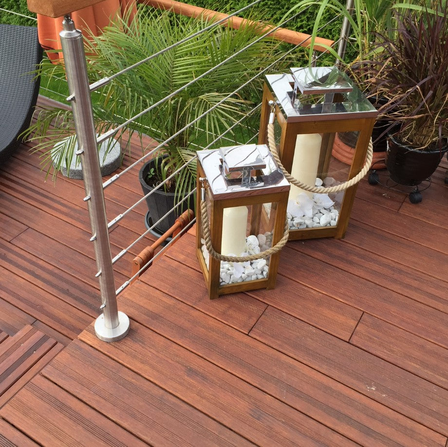 Detailaufnahme einer Bambusterrasse aus rötlich-braunen Holzdielen mit moderner Edelstahlseil-Brüstung. Zwei dekorative Holzlaternen mit Kerzen und weißen Ziersteinen stehen zwischen verschiedenen Kübelpflanzen und verleihen dem Außenbereich eine stilvolle, mediterrane Atmosphäre.