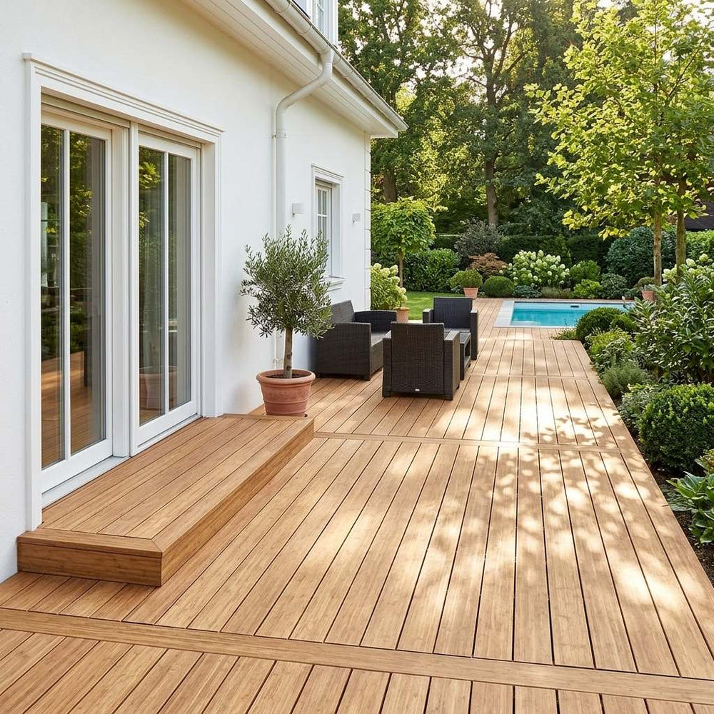 Weitläufige AMBOOO-Terrasse mit Sitzgruppe und Olivenbaum vor einem weißen Wohnhaus, mit Blick auf einen Pool und einen grünen Garten.