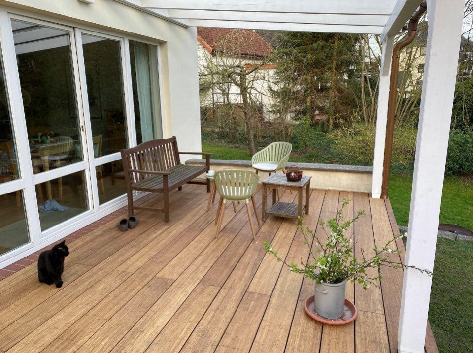 Moderne Terrasse mit hochwertigen glatten XL Bambus Terrassendielen, in der Farbe Sand  unter einer weißen Pergola. Gemütlicher Sitzbereich mit Gartenblick direkt am Wohnhaus mit großer Fensterfront.