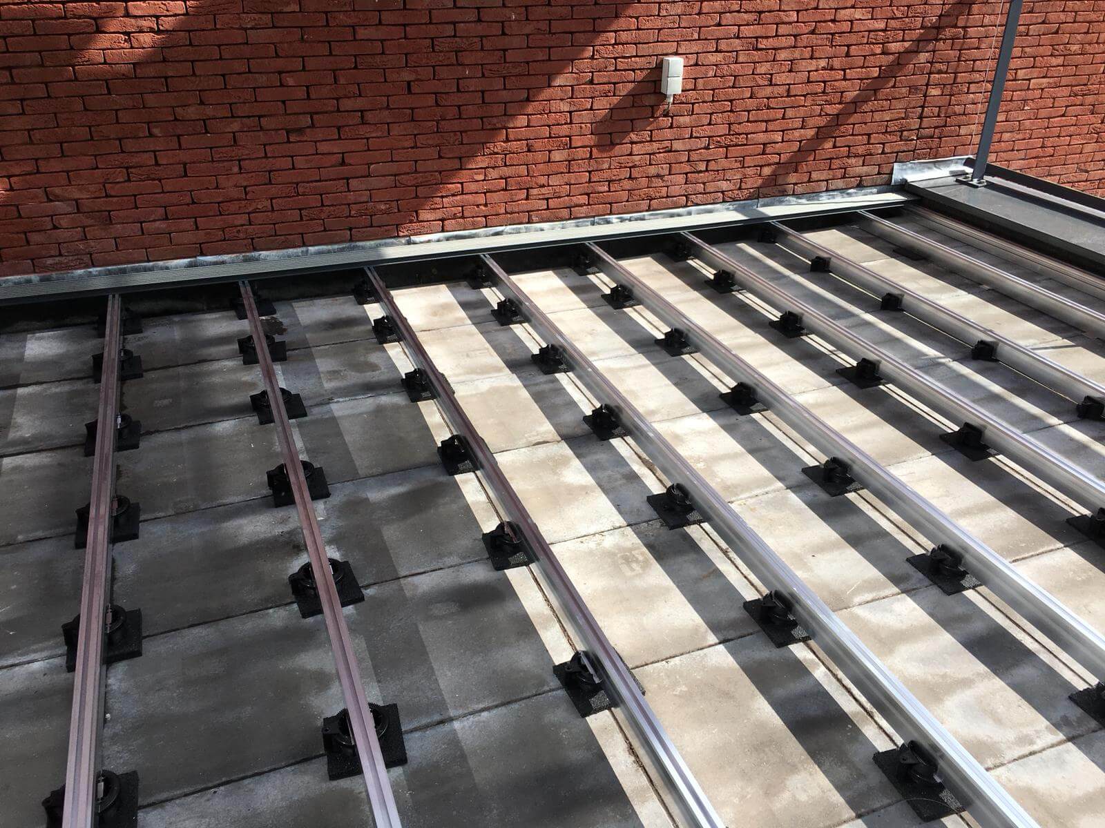 Aluminium-Streben als Unterkonstruktion für einen Terrassenbelag sind auf höhenverstellbaren Stelzlagern über Terrassenplatten montiert. Im Hintergrund ist eine Klinkerfassade zu sehen.