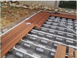 Eine Aluminium-Unterkonstruktion für eine Terrasse ist auf Betonsteinen verlegt. Ein Teil der braunen Terrassendielen wurde bereits montiert, während die restliche Unterkonstruktion für die weitere Verlegung vorbereitet ist.