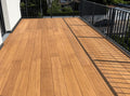 Großzügige Balkonterrasse, erstellt mit den AMBOOO Closed-Decking Dielen. Die Terrassendielen sind längs verlegt und werden von einem modernen anthrazitfarbenen Metallgeländer eingefasst. Sonnenschein erzeugt markante Schattenlinien des Geländers auf dem Terrassenboden. Im Hintergrund sind Wohnhäuser und Grünflächen zu sehen.
