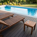Zwei Holzliegen und ein kleiner Beistelltisch stehen auf einer dunklen Poolterrasse mit Blick auf einen rechteckigen Swimmingpool und einen grünen Garten.