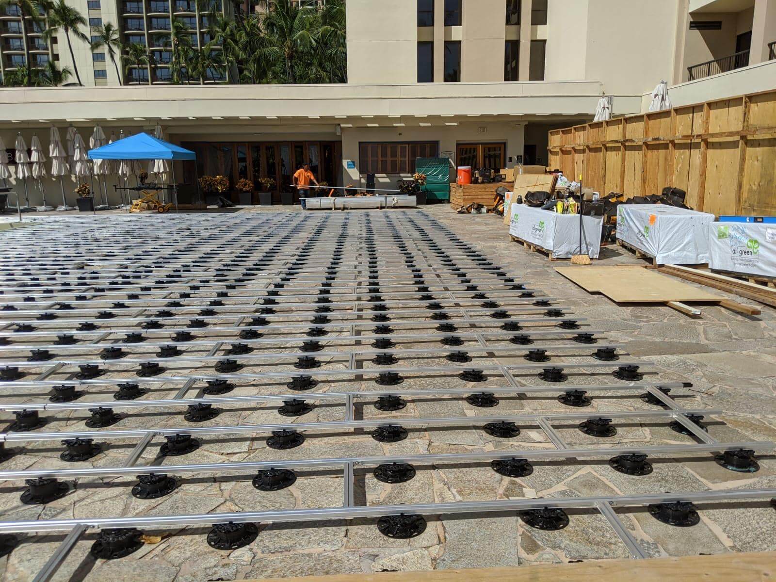 Großflächig verlegte Aluminium-Unterkonstruktion für eine Terrassenanlage. Die Aluminium-Unterkonstruktionen sind auf höhenverstellbaren Stelzlagern montiert; im Hintergrund befinden sich Gebäude, Palmen sowie Material und Werkzeuge für die Installation.