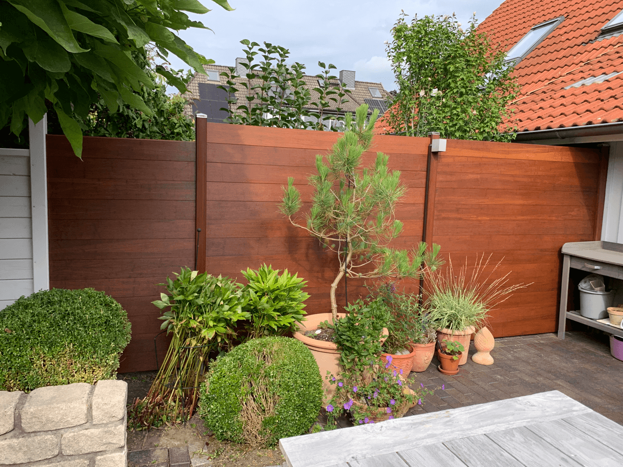 Moderner Sichtschutzzaun in warmer Bambusoptik mit dunklen Pfosten, der eine Terrasse begrenzt und als Hintergrund für diverse Gartenpflanzen und einen Pflanztisch dient.