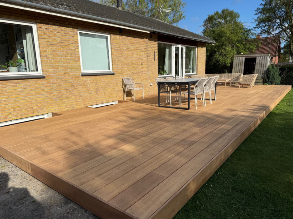 Großzügige Terrasse aus aMBOOO Closed Decking Bambus Terrassendielen in natürlicher Holzoptik, ausgestattet mit modernem Essbereich und Lounge-Möbeln, vor einem Einfamilienhaus mit gelber Klinkerfassade – pflegeleicht und wetterbeständig.