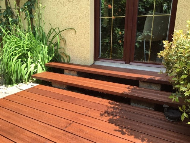 Terrasse mit rötlich-braunen Bambus-Terrassendielen und zwei passenden Bambus-Stufen vor einer Terrassentür, umgeben von Ziergräsern und Sträuchern.
