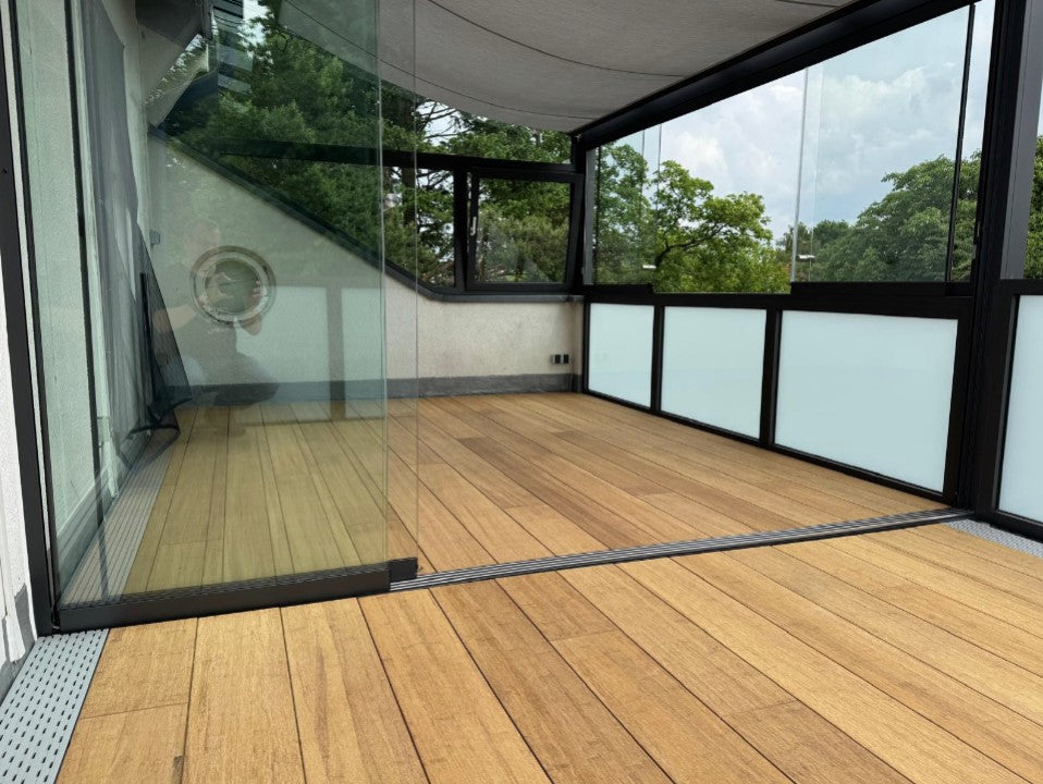 Moderner, verglaster Balkon mit hochwertigem Closed Deck Terrassenbelag in natürlicher Bambusoptik. Die bodentiefen Glasschiebeelemente und die dunklen Aluminiumrahmen schaffen einen geschützten Außenbereich mit freiem Blick ins Grüne. Der warme Braunton des Bodenbelags bildet einen harmonischen Kontrast zu den klaren Linien der Konstruktion und sorgt für eine elegante, zeitgemäße Atmosphäre.