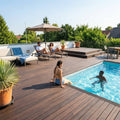 Großzügige Poolterrasse mit dunkelbraunen Terrassendielen, Sonnenliegen, Sitzbereich und Menschen beim Entspannen am Pool.