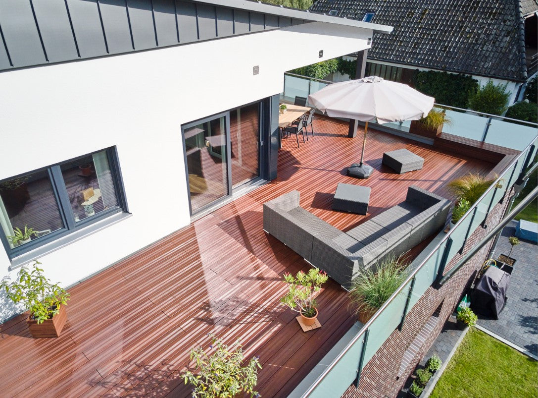 Moderne Dachterrasse mit edlen Bambus-Terrassendielen, gemütlicher Sitzlandschaft und Sonnenschirm. Das Glasgeländer bietet einen freien Blick auf die umliegenden Gärten und Häuser.