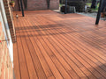 Großzügige Holzterrasse aus rötlich-braunen Terrassendielen an einer Backsteinfassade, teilweise überdacht von einer modernen Pergola mit dunklen Pfosten. Die Dielen sind längs verlegt und wirken gepflegt, während Sonnenlicht und Schattenmuster über die Fläche fallen. Im Hintergrund befinden sich weitere Backsteinwände, Pflanzgefäße und ein Gartenbereich.
